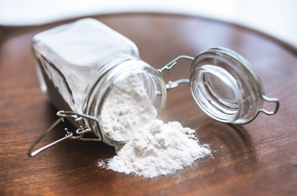 Ein umgekipptes, geöffnetes Bügelglas liegt auf einer Holzoberfläche, sodass weißes Backpulver oder Mehl aus der Öffnung herausrieselt.