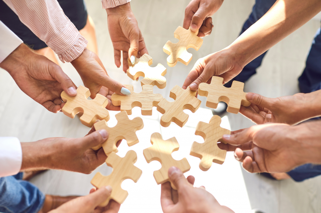 Mehrere Hände halten im Kreis große Puzzleteile aus Holz zusammen, die symbolisch für Teamwork und Zusammenarbeit stehen.