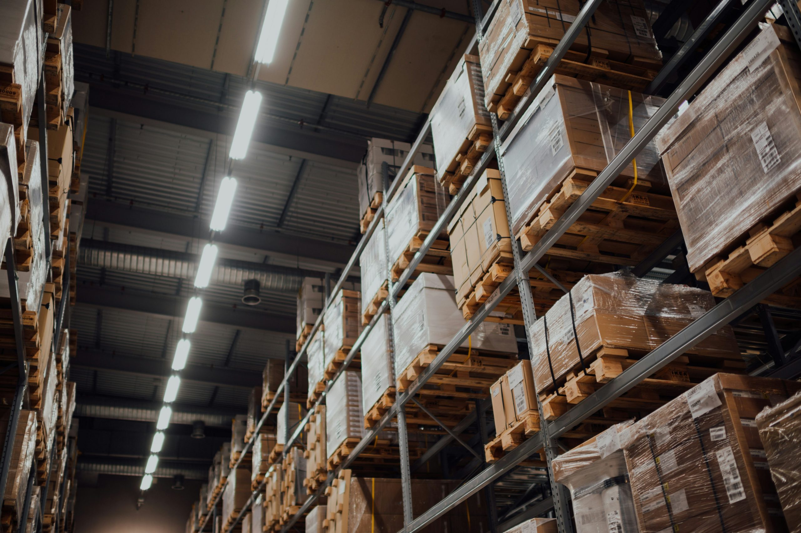 In die Höhe ragende, industrielle Schwerlastregale in einer Lagerhalle, gefüllt mit in Folie verpackten Paletten, die Kisten und Waren enthalten, beleuchtet durch eine Reihe von Neonröhren an der Decke.